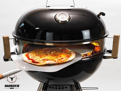 Pizza-Aufsatz auf schwarzem Kugelgrill mit frischer Pizza auf Schaufel